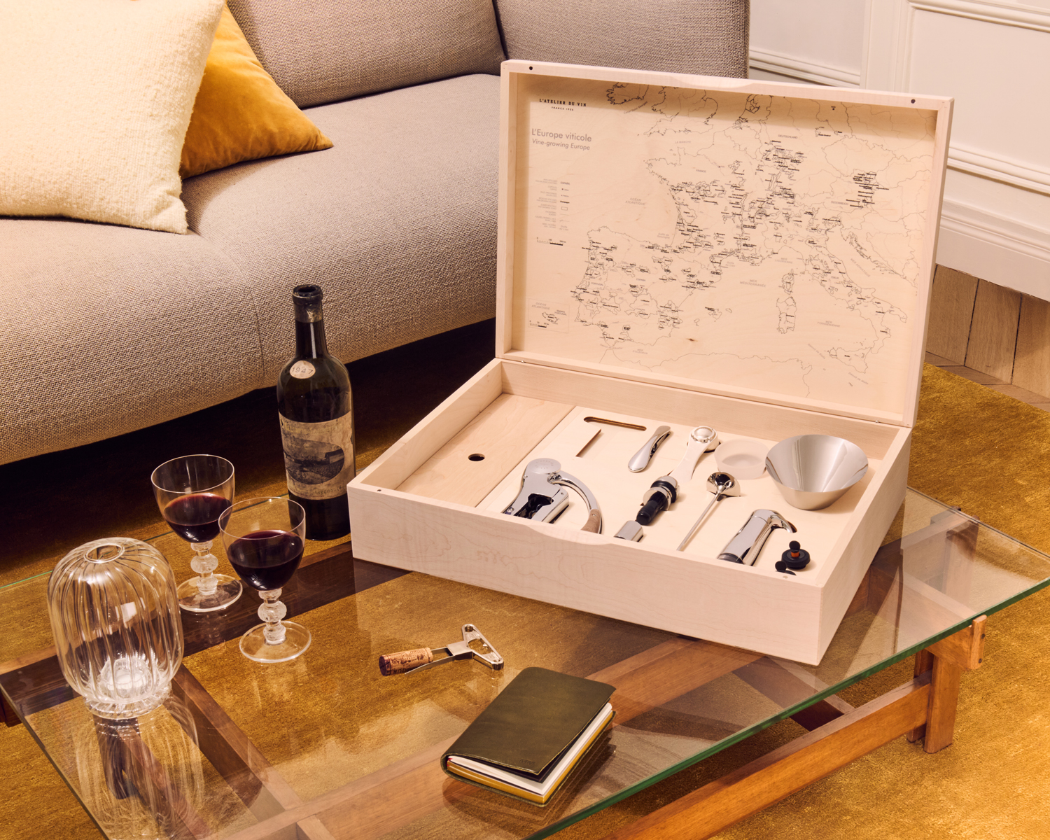 Coffret œnologique en bois de L'Atelier du Vin avec de nombreux Outils du Vin posé sur une table de salon avec une bouteille de vin rouge 